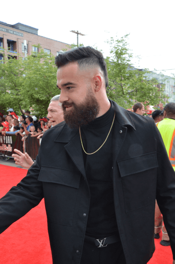 Carlos Coronel with a beard wearing a black outfit walks on a red carpet, surrounded by fans and greenery in the background entering Red Bull Arena after the red carpet team walk in.