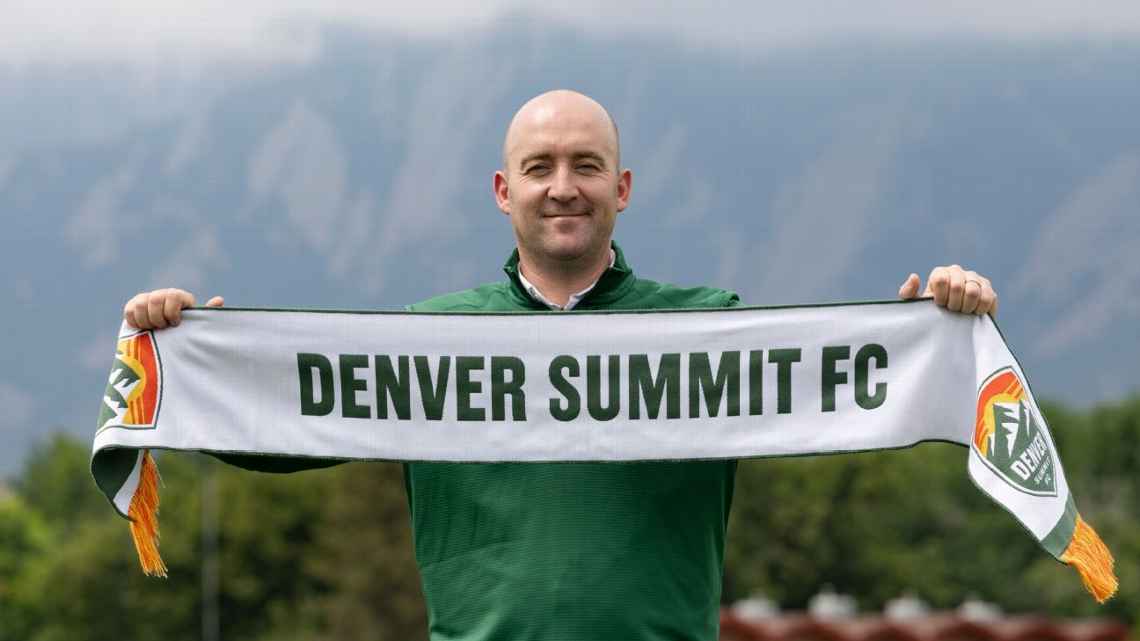 Nick Cushing holds Denver Summit FC scarf while wearing green jacket with mountains in background