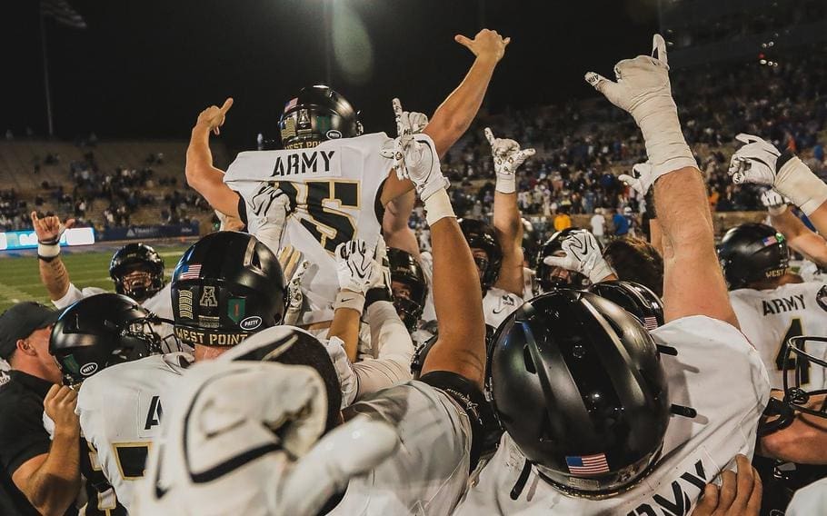 Army West Point football players clad in white away jerseys and black helmets lift their kicker Dawson Jones into the air in a jubilant celebration on the field at night following a game-winning field goal.