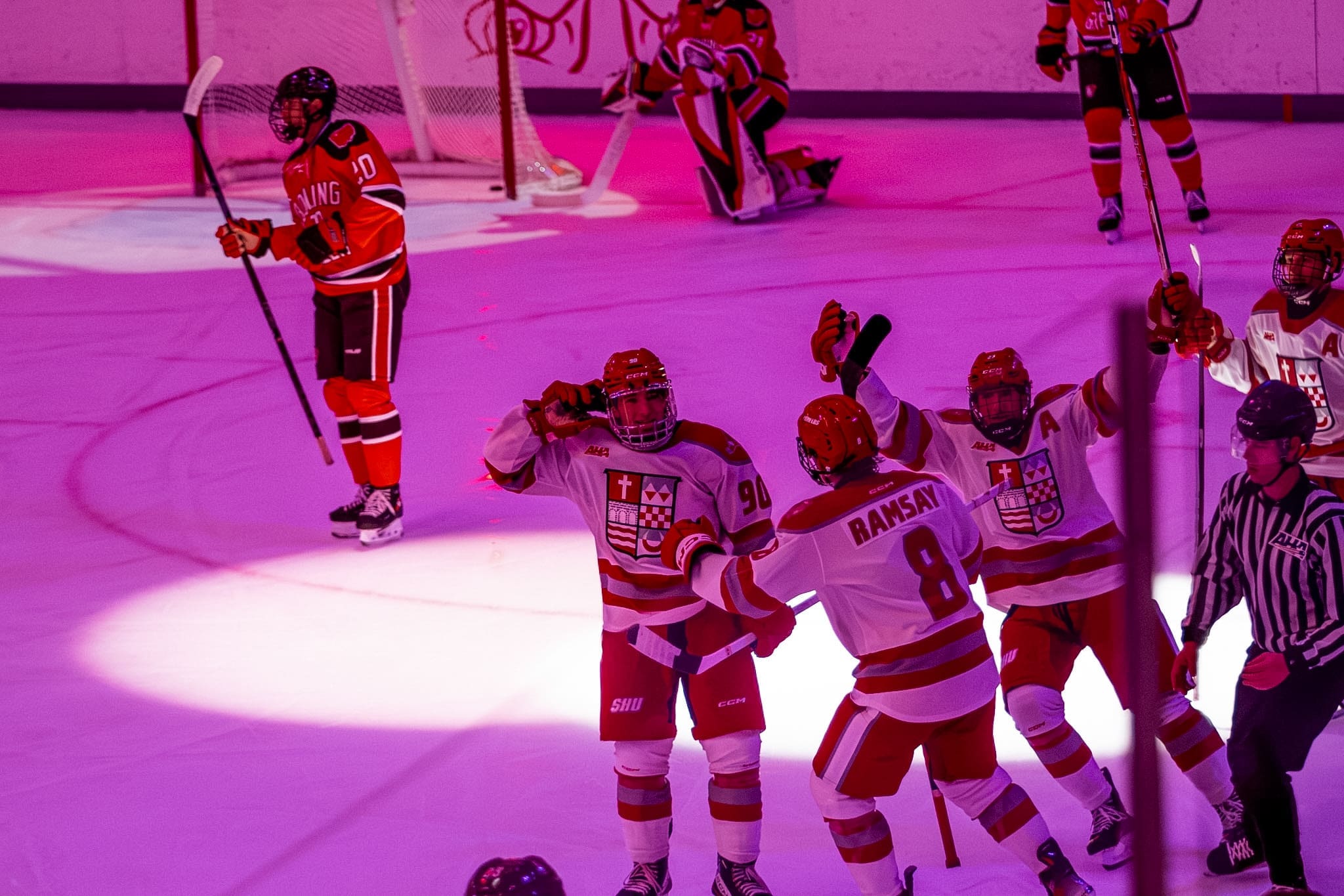 Sacred Heart Pioneers hockey forward Diego Buttazzoni wearing white jersey number ninety celebrates scoring a goal on the ice under purple arena lighting with teammates Chase Ramsay number eight and others while a Bowling Green player looks on in the background.