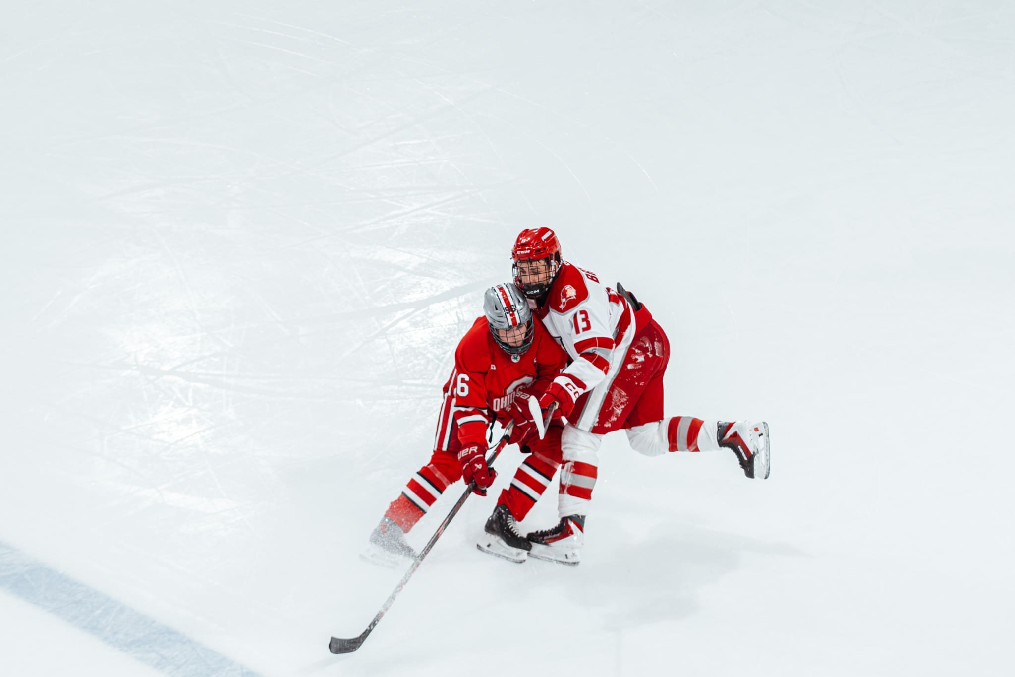Sacred Heart defenseman Nick Bernardo steps up at the blue line in a white and red jersey to deliver a heavy open-ice body check from behind on an Ohio State forward wearing a red jersey.