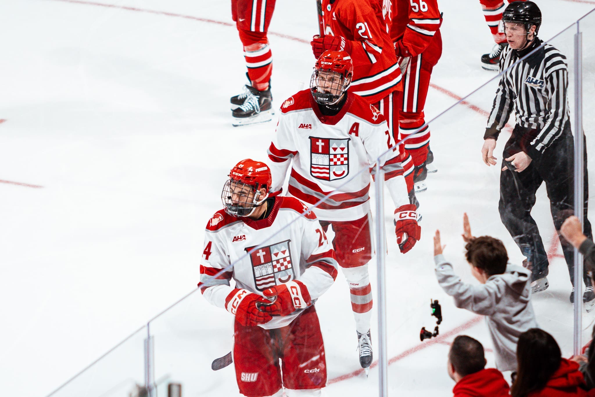 Sacred Heart defenseman Taos Jordan (4) skates past the home team bench while a teammate skates behind him and fans cheer along the glass.