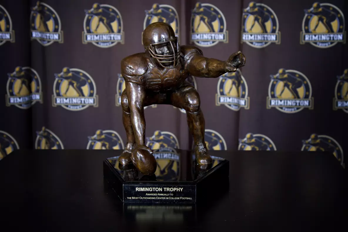 A bronze sculpture of a football center crouched over a football, mounted on a black base inscribed with "Rimington Trophy," set against a step-and-repeat backdrop featuring the award logo.