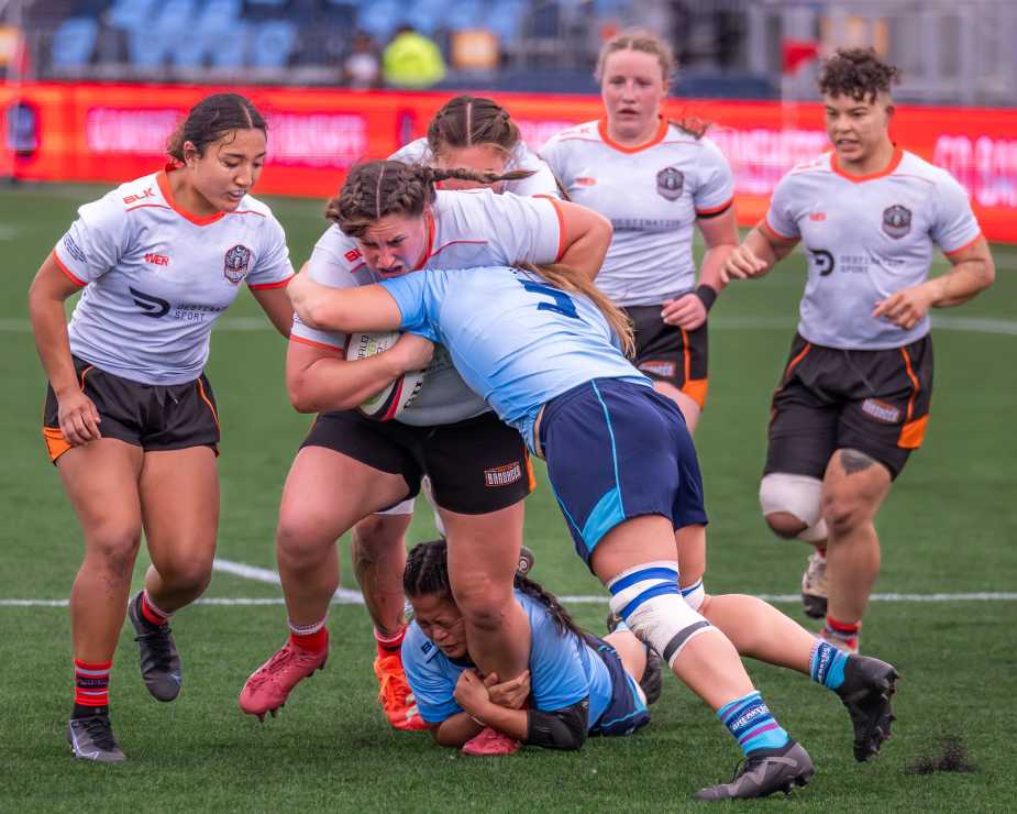 Two rugby players collide in a physical match, with one player in blue attempting to tackle another in a black and white jersey, while teammates surround them during a game.
