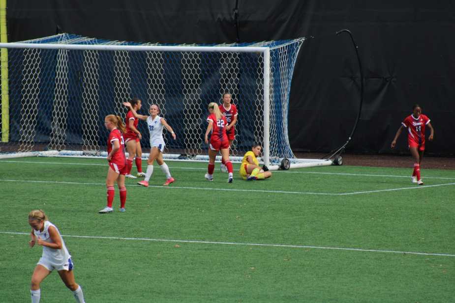 St. John's goalkeeper Kayla Bower in pink uniform makes a diving save in a crowded penalty area with multiple Seton Hall players in red and St. John's defenders in white positioned near the goal