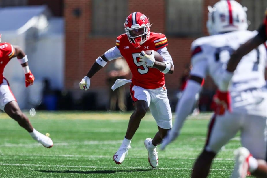 Maryland football players in red and white uniforms celebrate together in the end zone after scoring a touchdown, with teammates gathering around and pointing in celebration at SECU Stadium