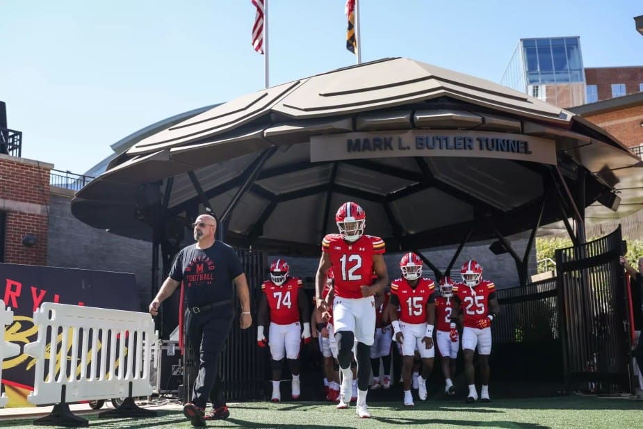 Maryland football team emerges from the Mark L. Butler Tunnel at SECU Stadium, led by quarterback Jayden Shipps wearing number 12, with players in red and white uniforms ready for pre-game warmups