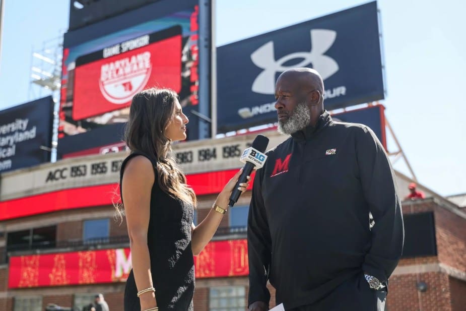 Maryland head coach Michael Locksley conducts a pre-game interview with a female reporter holding a Big Ten Network microphone, with SECU Stadium and Under Armour signage visible in the background