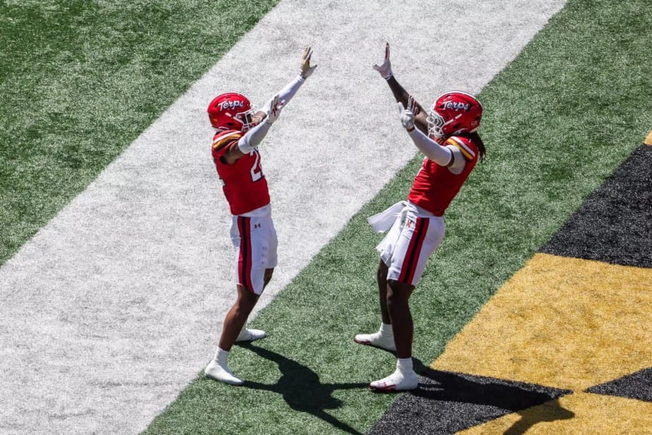 Two Maryland football players in red uniforms celebrate together in the end zone, both pointing upward with their arms raised in triumph