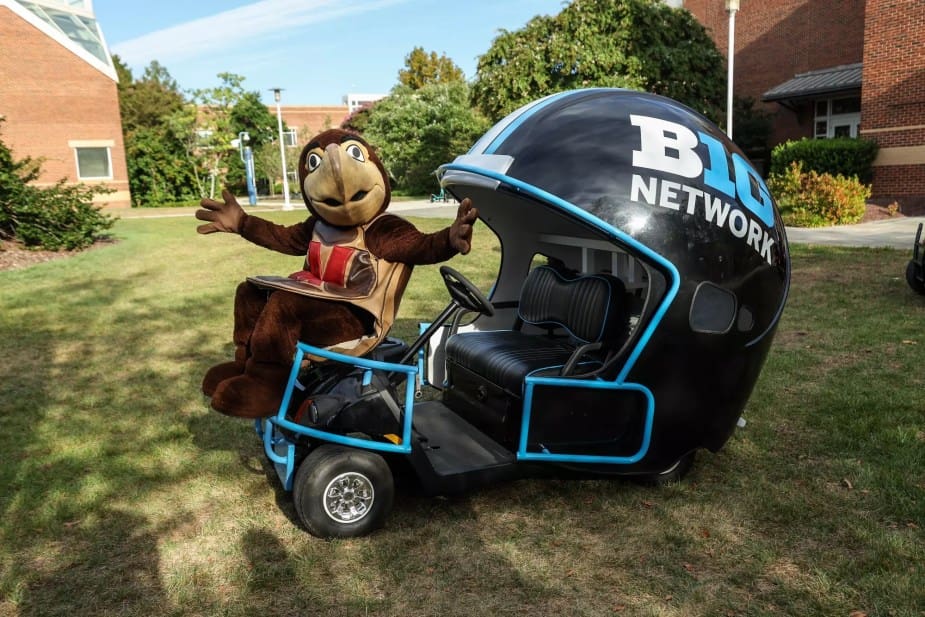 Maryland Terrapins mascot Testudo the turtle sits in a Big Ten Network branded golf cart on a college campus, with arms raised in celebration and brick buildings visible in the background